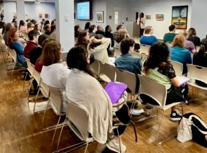 College professor talking at podium in a large room with people sitting listening to her.