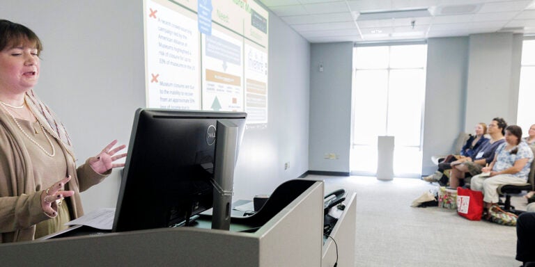 A business professional presents as she stands at a computer while talking to conference guests sitting in the room with a projector screen.