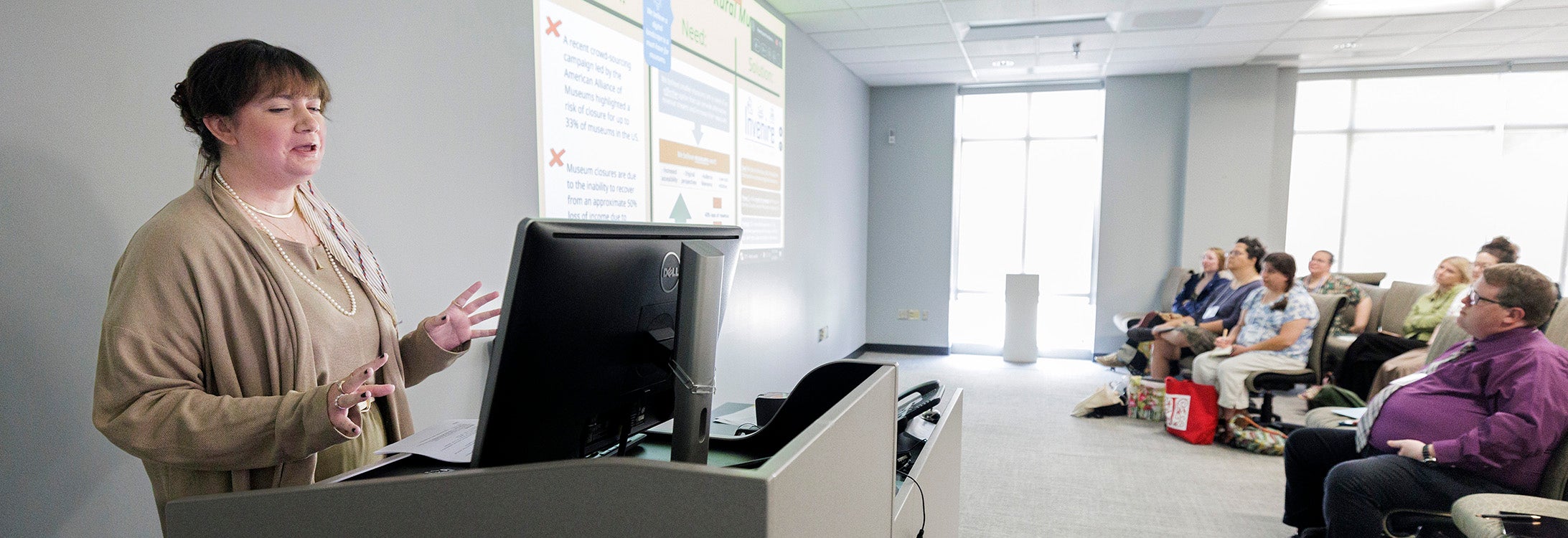 A business professional presents as she stands at a computer while talking to conference guests sitting in the room with a projector screen.